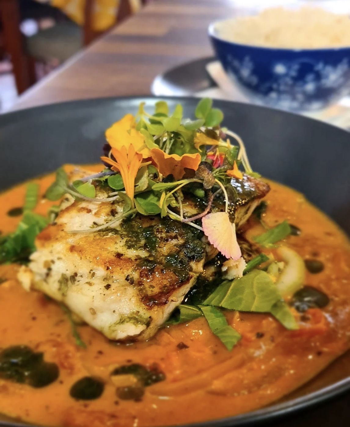Grilled fish in an orange curry with herbs and edible flowers, rice in a blue bowl behind